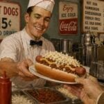 A traditional West Virginia hot dog topped with mustard, chili, onions, and coleslaw is served in a 1950s diner setting. The chili-slaw dog became a popular meal among miners, railroad workers, and factory employees and remains a signature food across the Mountain State. (WVExplorer Illustration)
