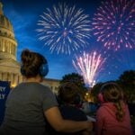 A family watches an Independence Day fireworks display from a designated sensory-friendly viewing area while wearing hearing protection. More communities are adding quiet spaces, ear protection, and other accommodations to help people with autism, PTSD, and sensory sensitivities enjoy public celebrations. (WVExplorer Illustration)