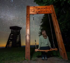 The “Almost Heaven” swing at Droop Mountain Battlefield State Park overlooks the mountains beneath a star-filled sky, with the park’s observation tower at left. The Pocahontas County battlefield has joined the West Virginia Paranormal Trail for 2026.