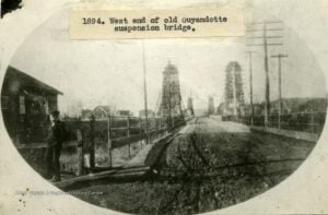 Historic 1894 photograph of the west end of the Guyandotte Suspension Bridge in Huntington, West Virginia, showing the bridge towers, roadway, wooden railings, and a young boy standing nearby.