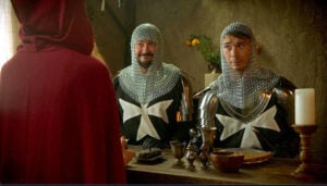 Two knights in chainmail and armor sit at a wooden table facing a hooded figure in a scene from The Kingdom of Rexmonte.