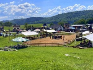 Medieval-style buildings, colorful tents, and a fenced arena at the West Virginia Renaissance Festival near Lewisburg, with forested mountains in the background.