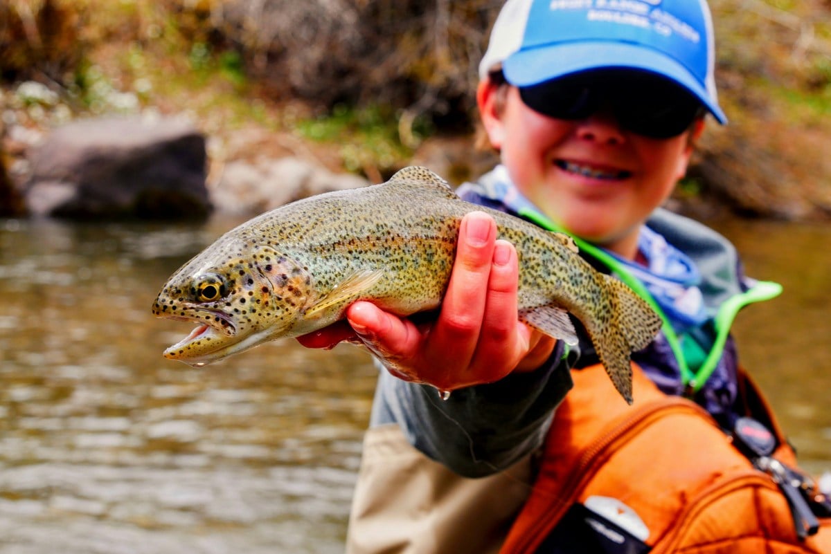 Young angler wearing a blue cap holds a large rainbow trout beside a rocky stream.