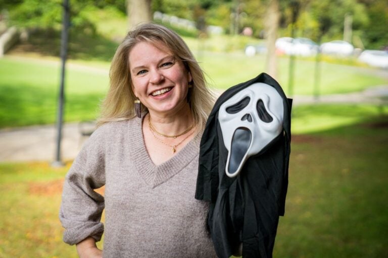 WVU professor Elizabeth Cohen smiles beside a Ghostface mask outdoors on campus.
