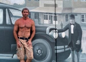 Artist Jesse Corlis stands shirtless in paint-stained shorts beside a detailed mural of a vintage automobile, historic building, and man in period clothing.