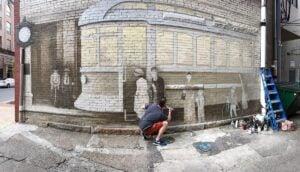 Artist Jesse Corlis crouches beside a brick building while painting a large mural depicting a historic streetscape.